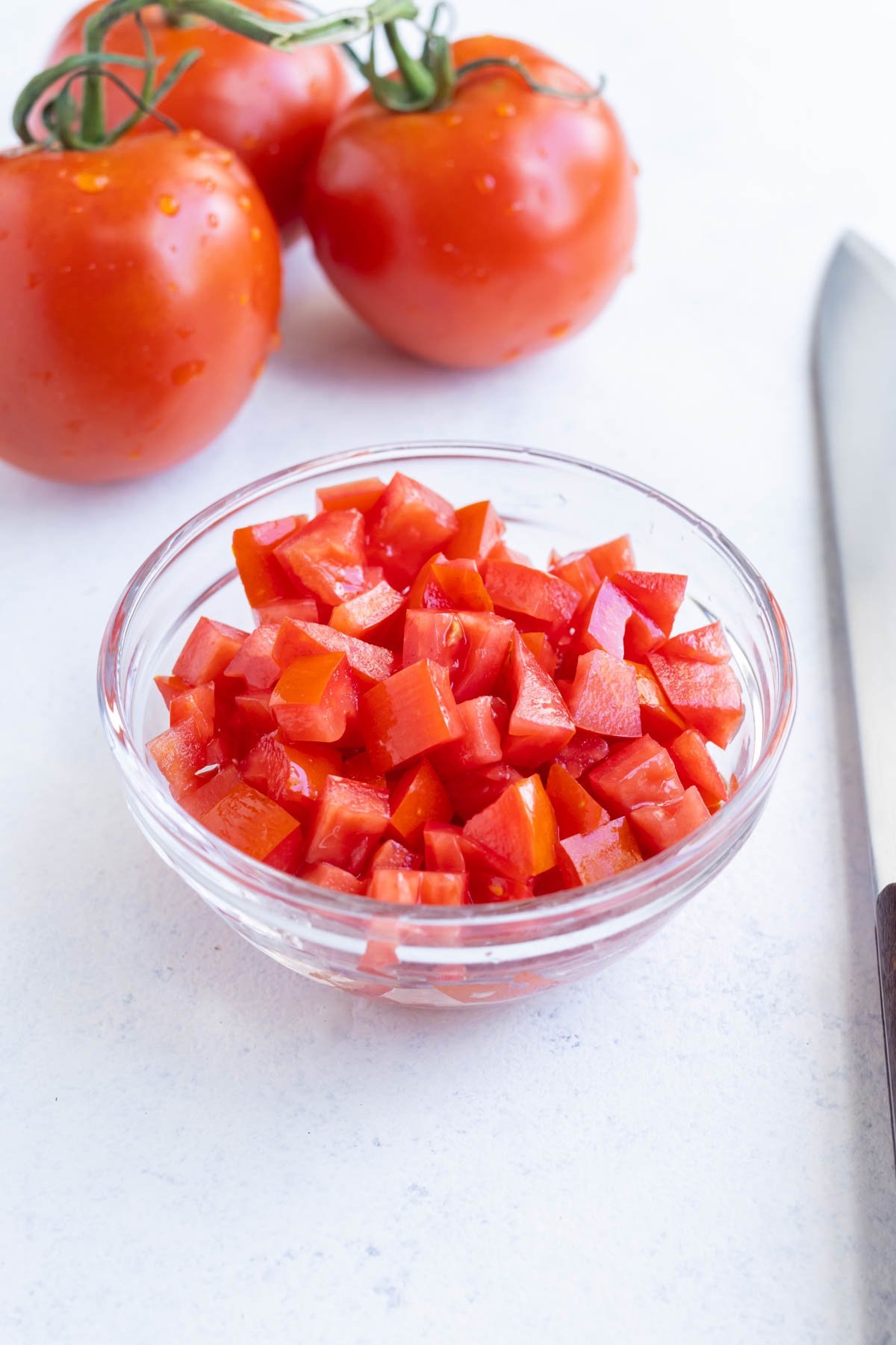 A small, glass bowl full of healthy diced tomatoes with whole tomatoes in the background.