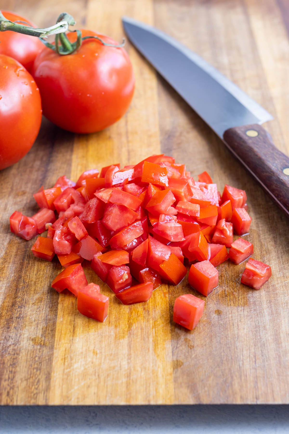 Diced tomato on a wooden cutting board with whole tomatoes and a sharp knife on the side.