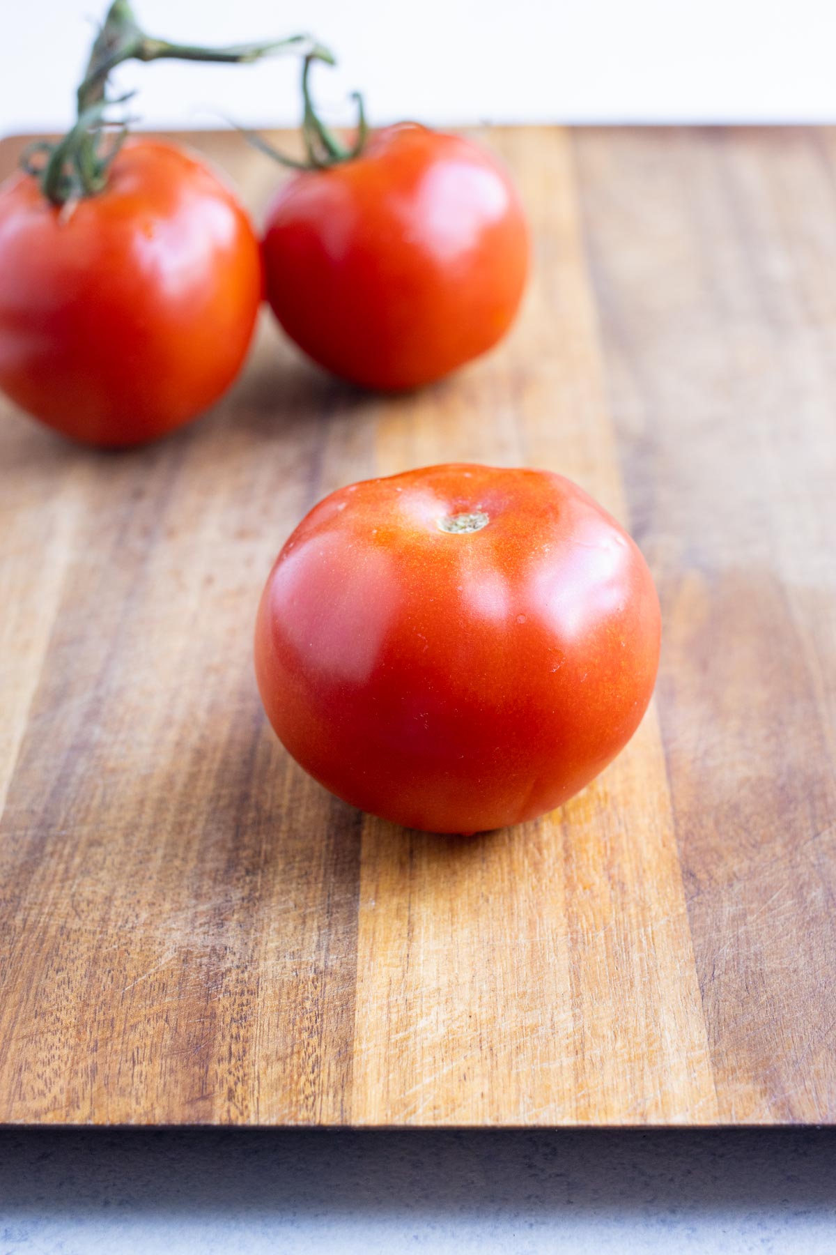 A firm, round tomato on a cutting board with other tomatoes on a vine in the background.