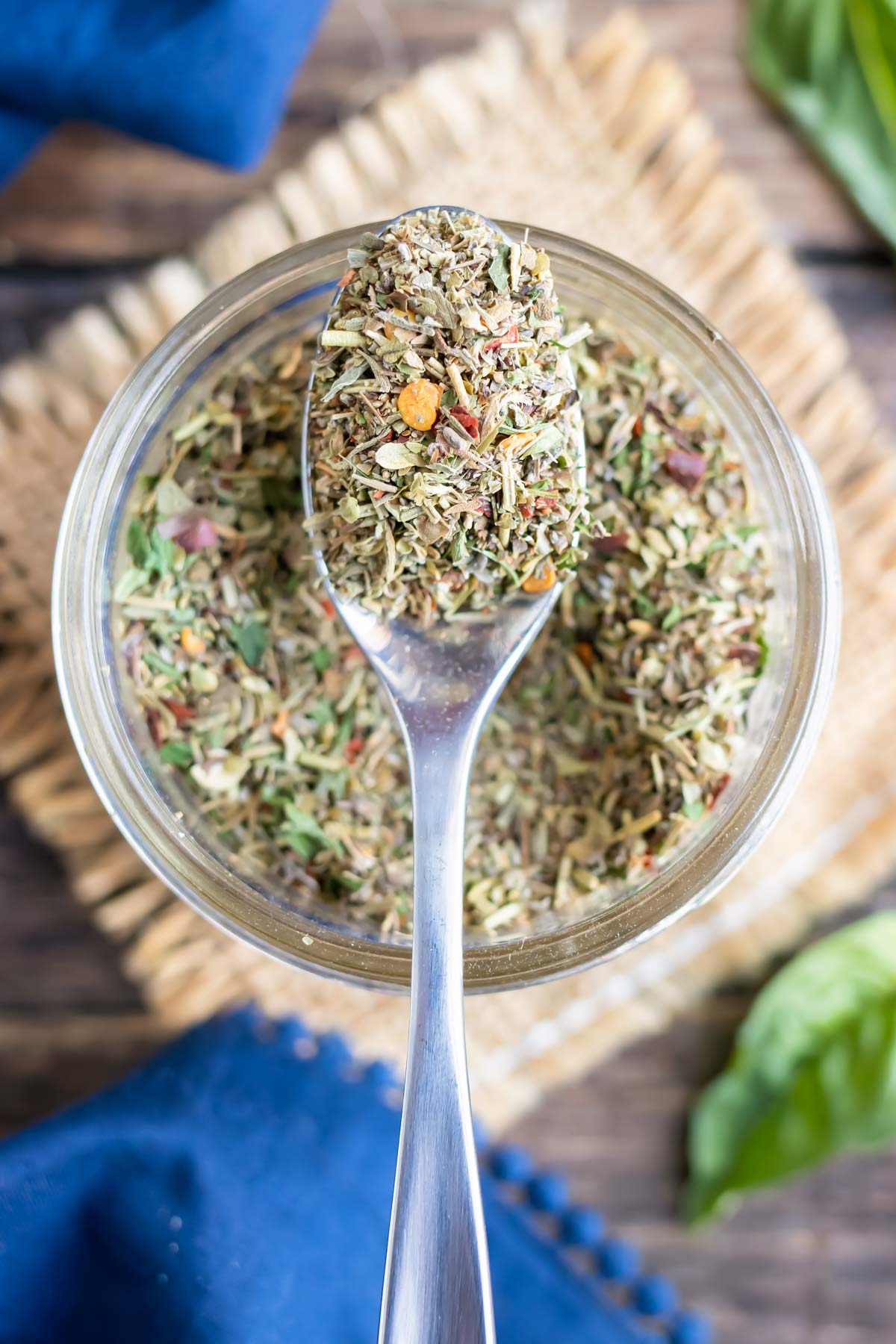 A clear glass jar with a spoon scooping out a DIY seasoning mix made with Italian herbs.