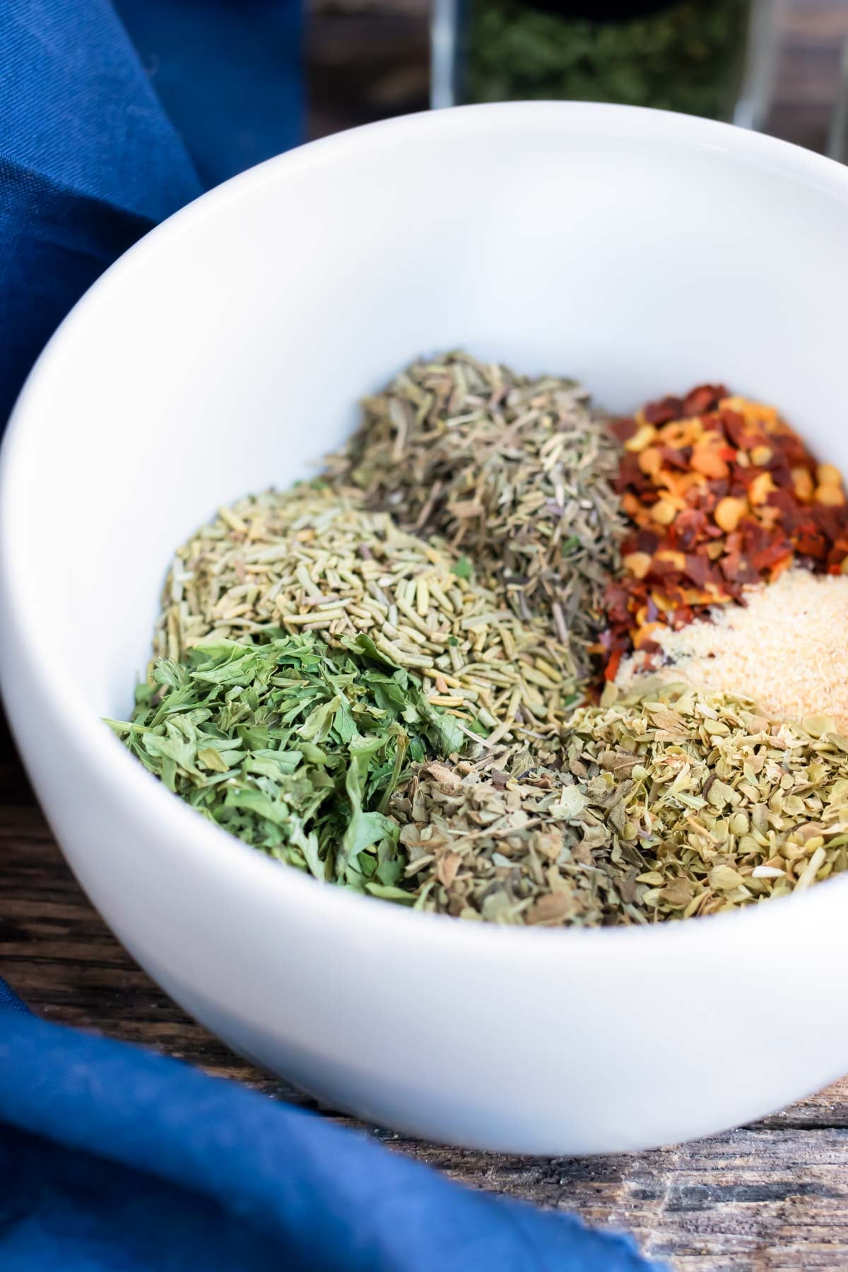 Dried herbs, garlic powder, and red pepper flakes in a white bowl before mixing Italian herb seasoning.