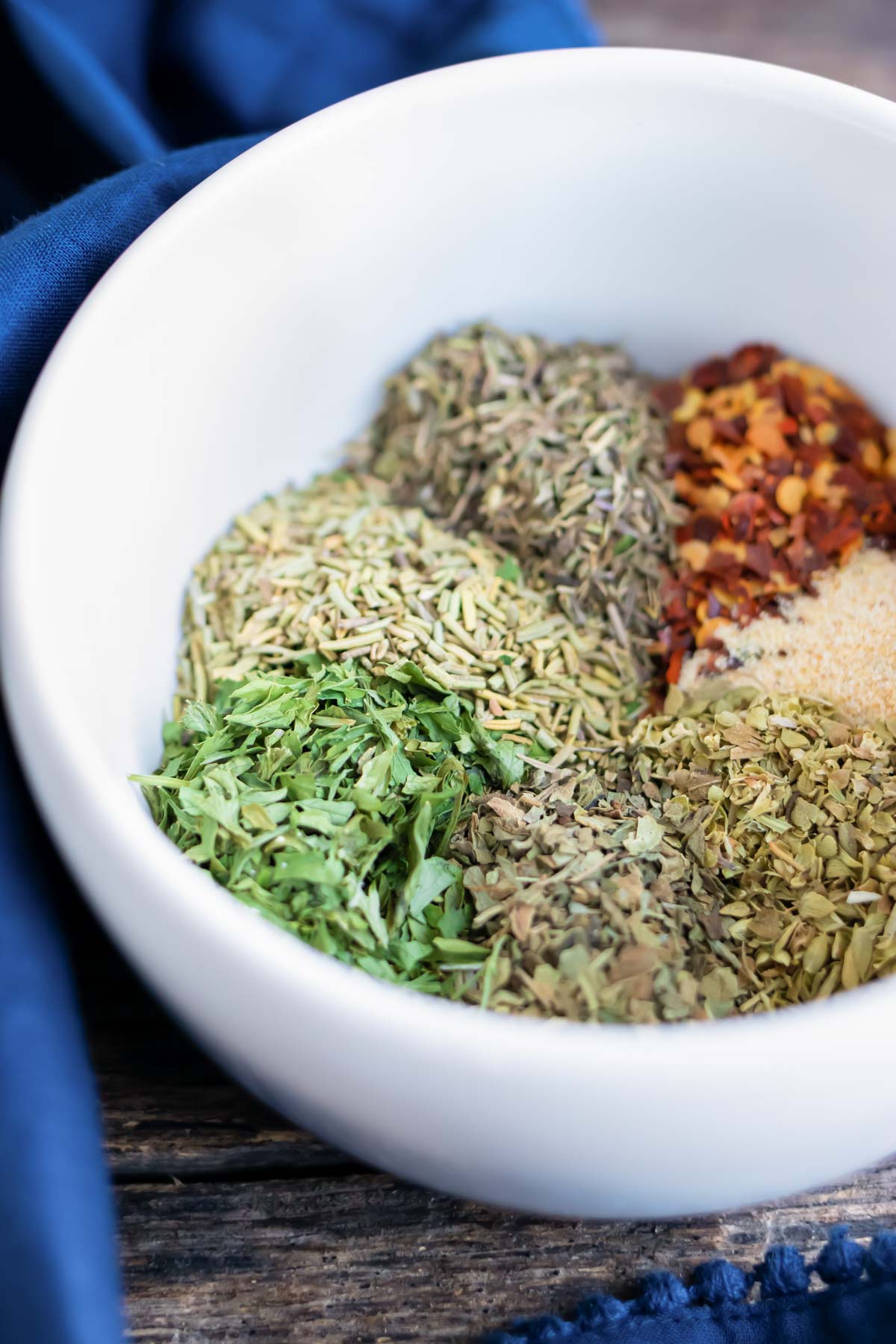 A white bowl full of dried oregano, basil, parsley, rosemary, thyme, and garlic powder for an Italian seasoning recipe.