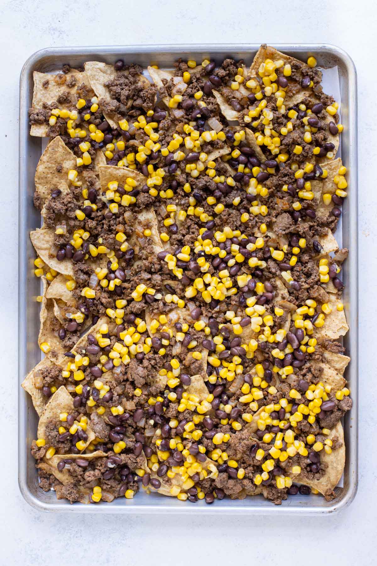Chips and ground beef are topped with corn and beans on a baking sheet.
