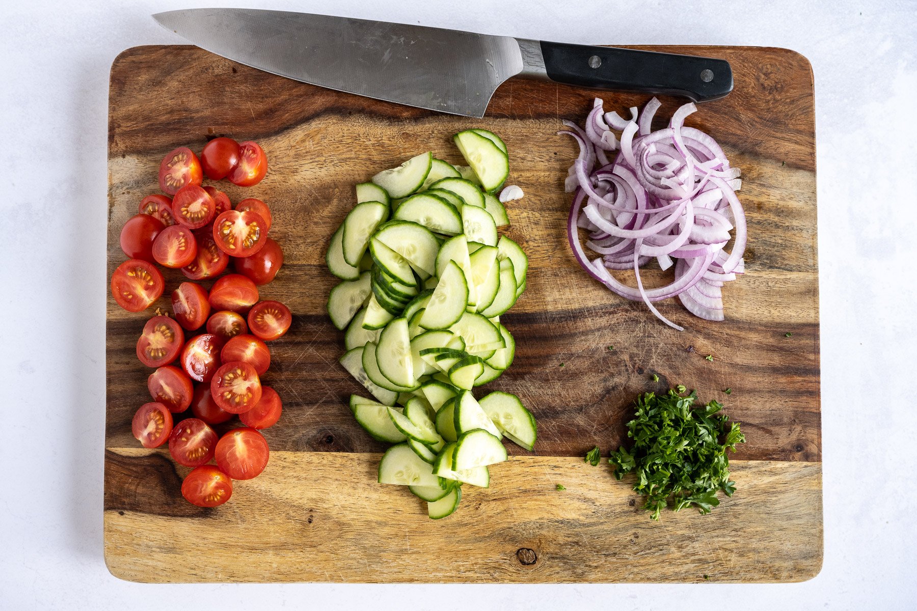 Chopped tomatoes, cucumbers, onions, and parsley are on a wooden cutting board.