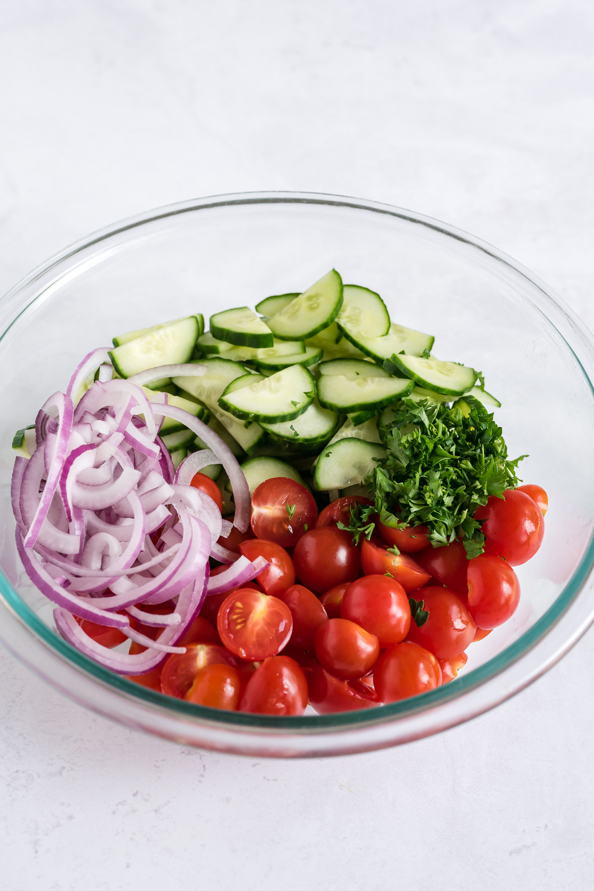 Sliced tomatoes, cucumbers, onions, and parsley in a glass bowl.