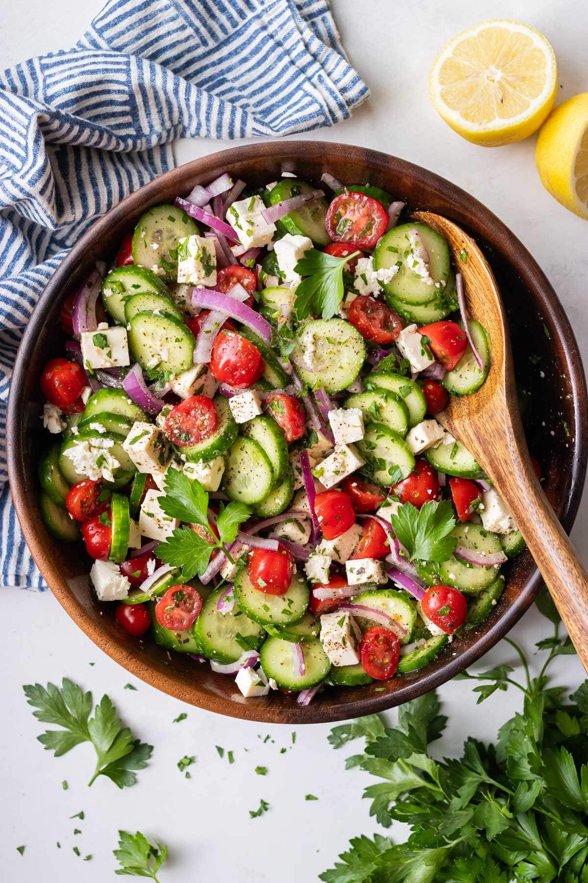 A Greek cucumber tomato and feta salad is served in a wooden bowl with a spoon and lemons next to it.