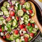 A Greek cucumber tomato and feta salad is served in a wooden bowl for a simple summer side dish.