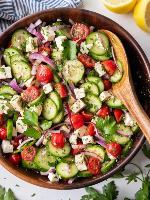 A Greek cucumber tomato and feta salad is served in a wooden bowl for a simple summer side dish.