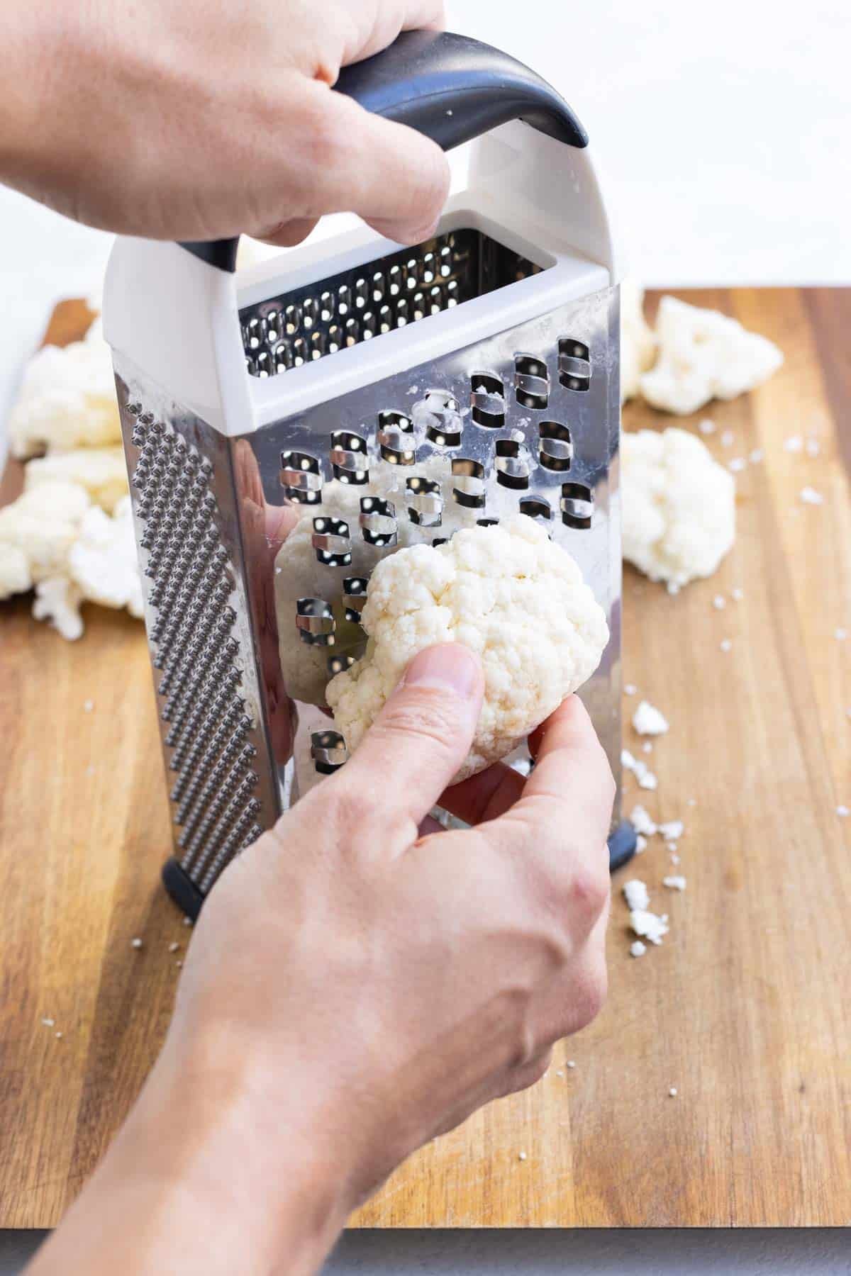 A hand carefully grates cauliflower on a cheese grater.