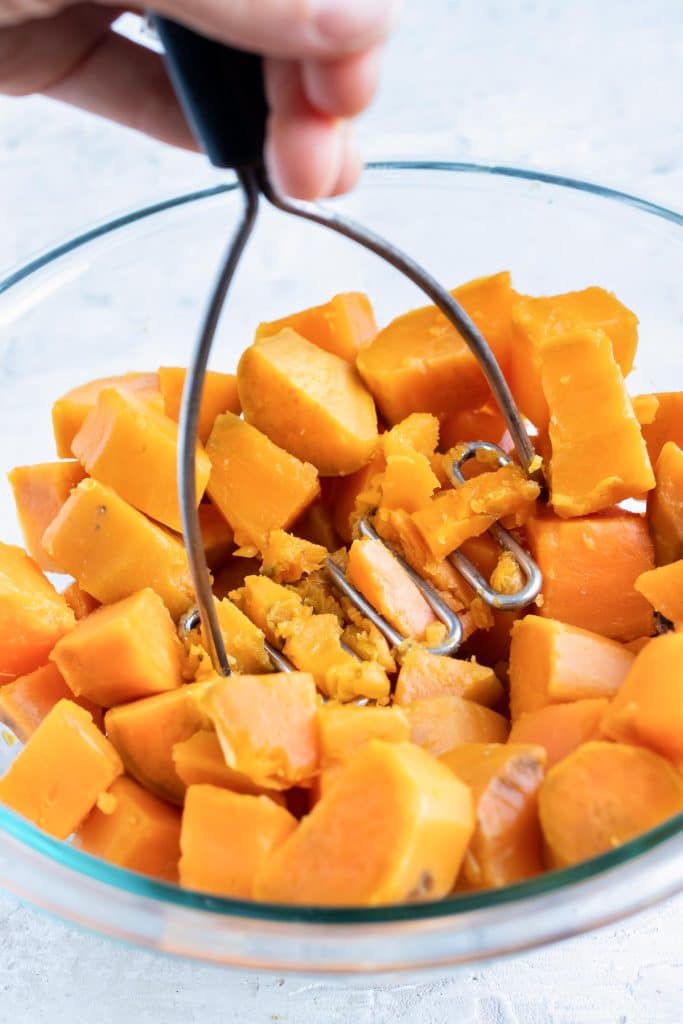A hand using a potato masher to mash sweet potatoes by hand in a glass bowl.