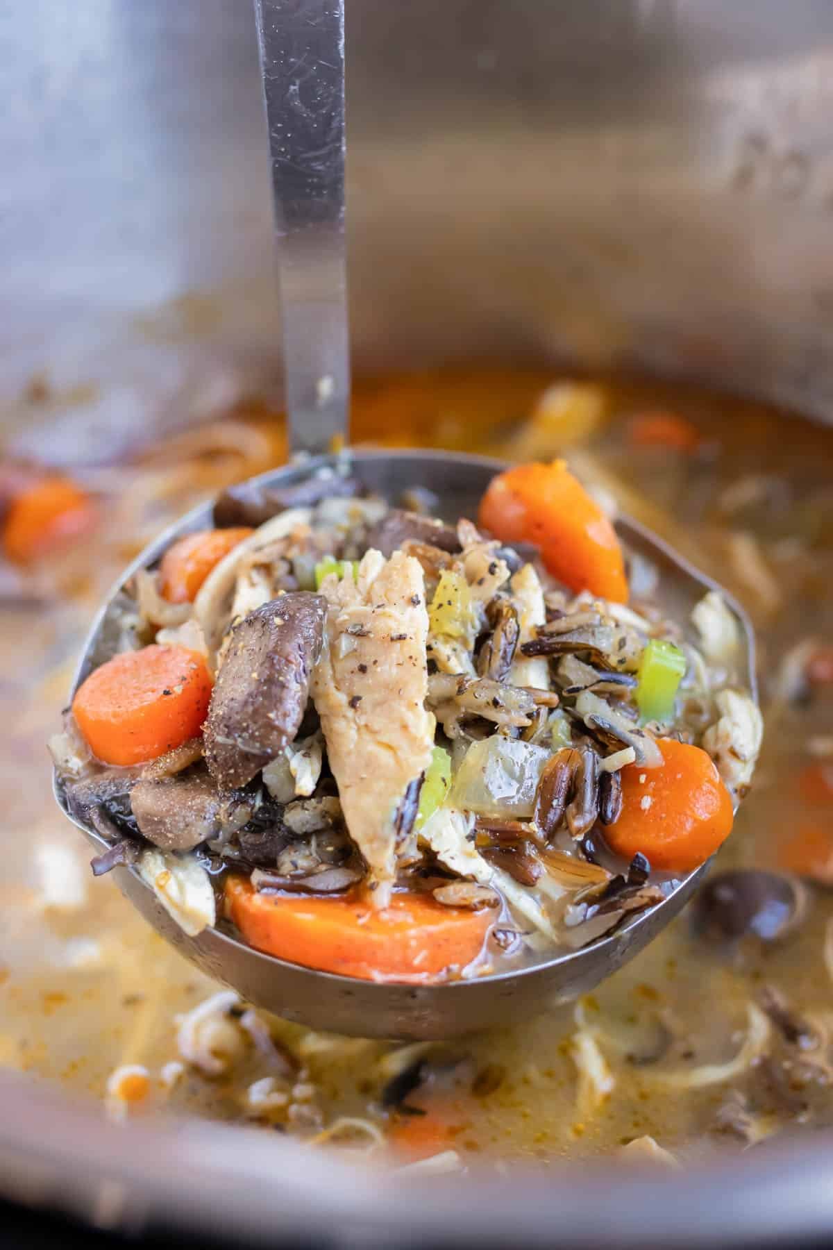 A ladle is full of chicken, mushrooms, vegetables, rice, and broth before serving.