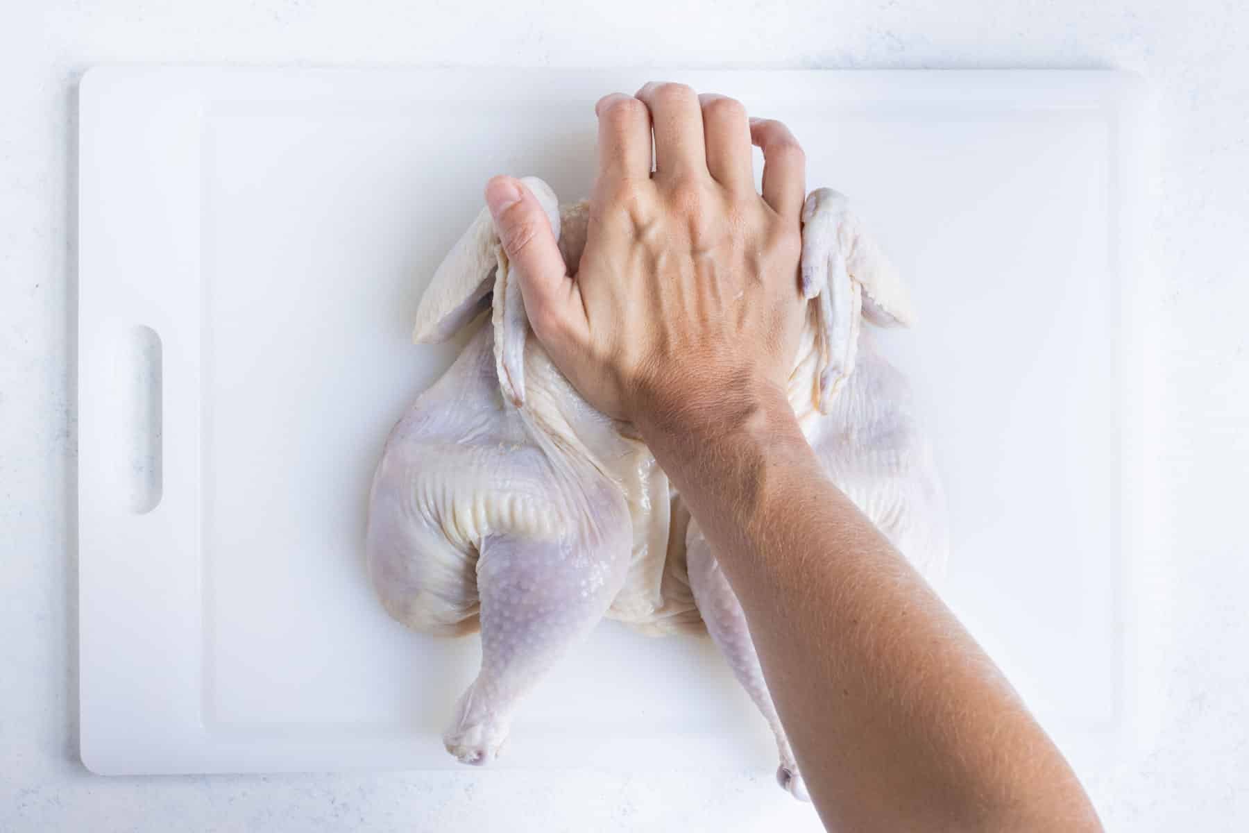 A hand presses down on a chicken with the spine removed to spatchcock it.