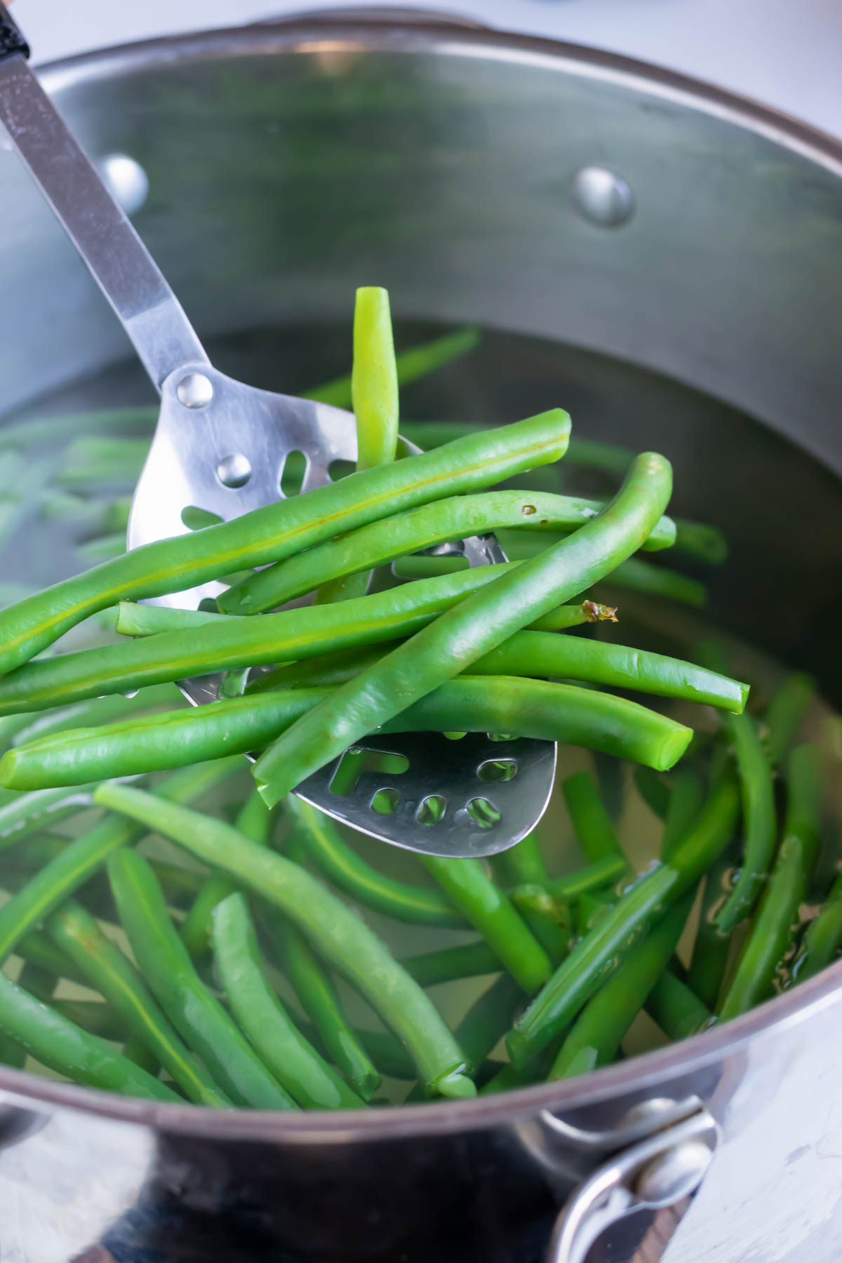 Boiled green beans being pulled out of a pot with a slotted spoon.