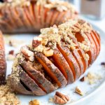 Hasselback sweet potato recipe is served on a white plate and topped with toasted pecans, brown sugar, and cinnamon.