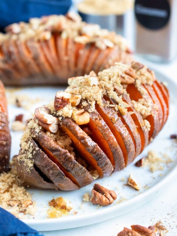 Hasselback sweet potato recipe is served on a white plate and topped with toasted pecans, brown sugar, and cinnamon.