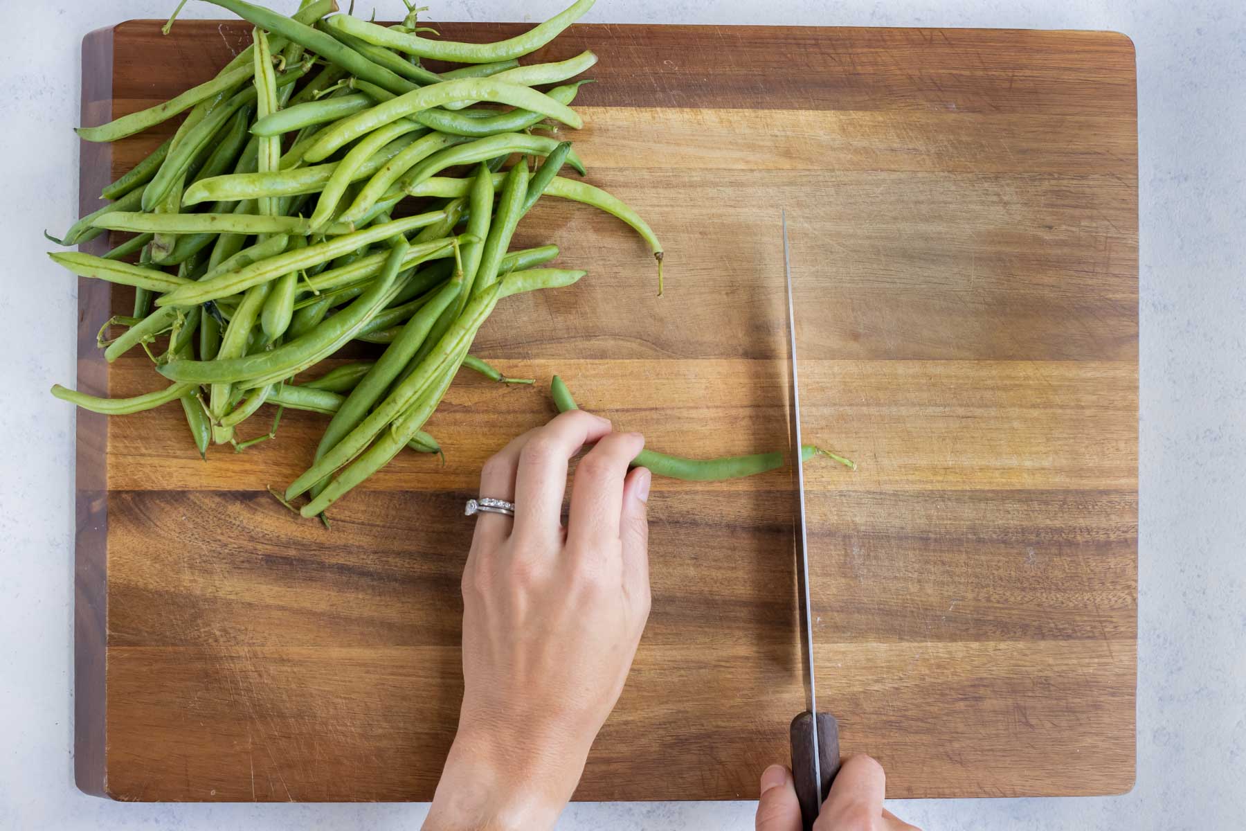 A green bean is cut with a knife on a cutting board.