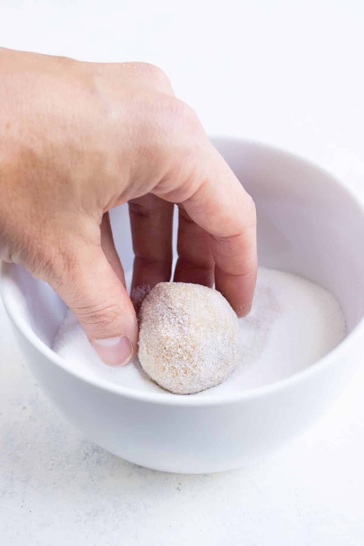 A hand rolls a peanut butter cookie dough ball in a bowl of sugar.