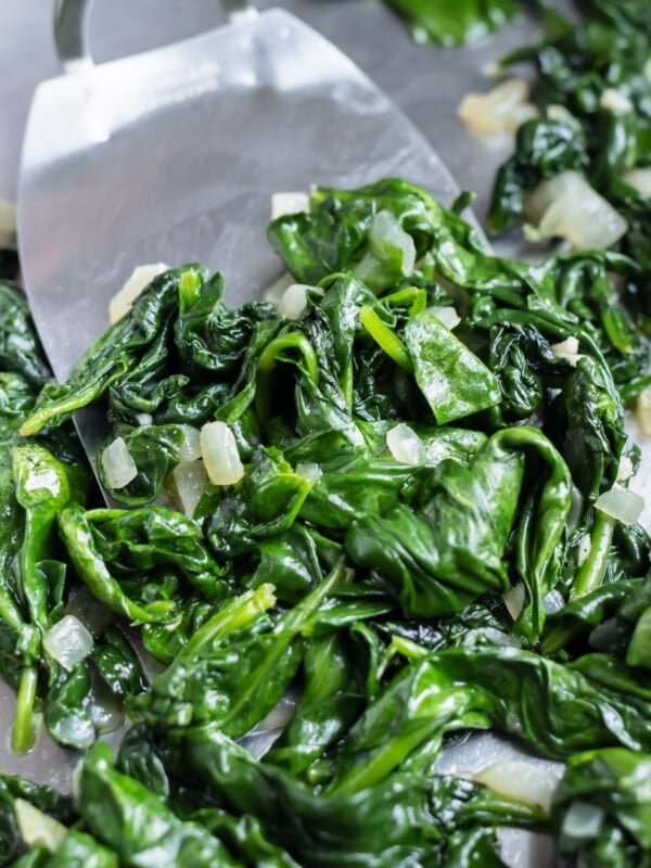 A spatula scooped up sauteed spinach with onions and garlic from a skillet.