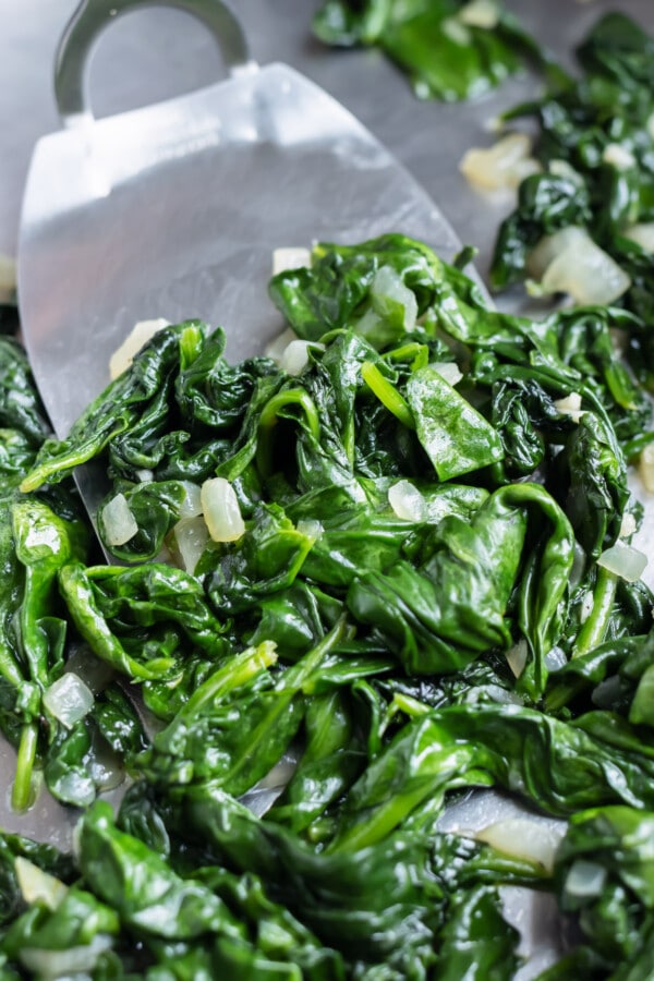 A spatula scooped up sauteed spinach with onions and garlic from a skillet.