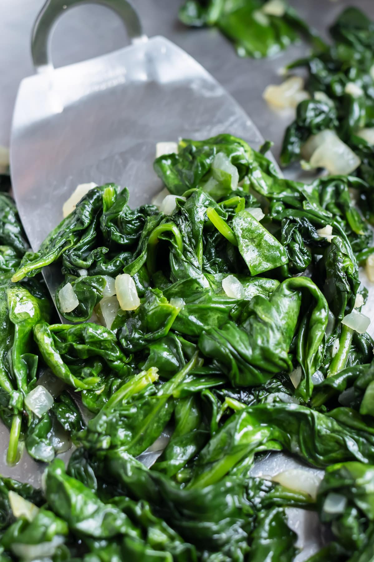 Sauteed Spinach RECIPE in a stovetop pan with a silver spatula about to scoop out some spinach.