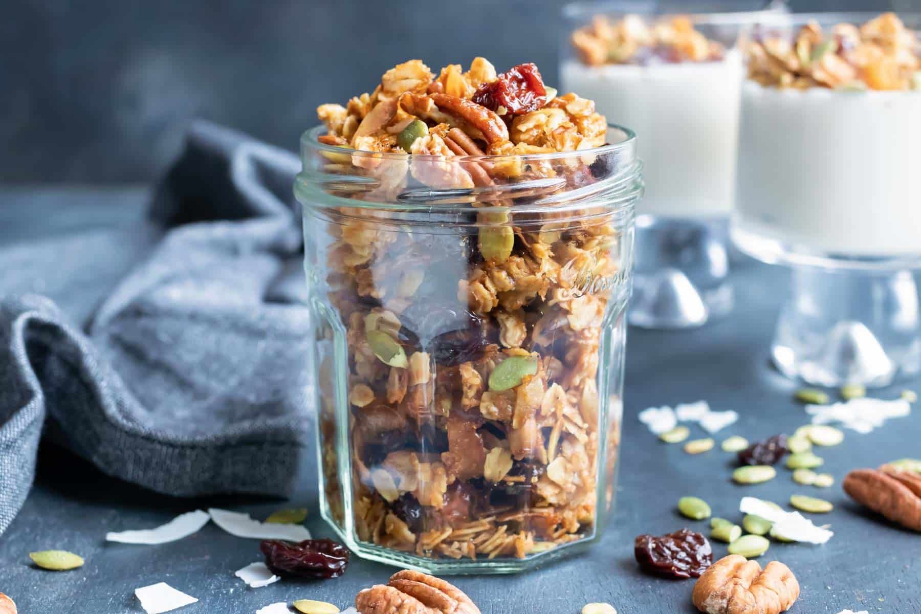 A mason jar full of homemade granola with two yogurt parfaits behind it.