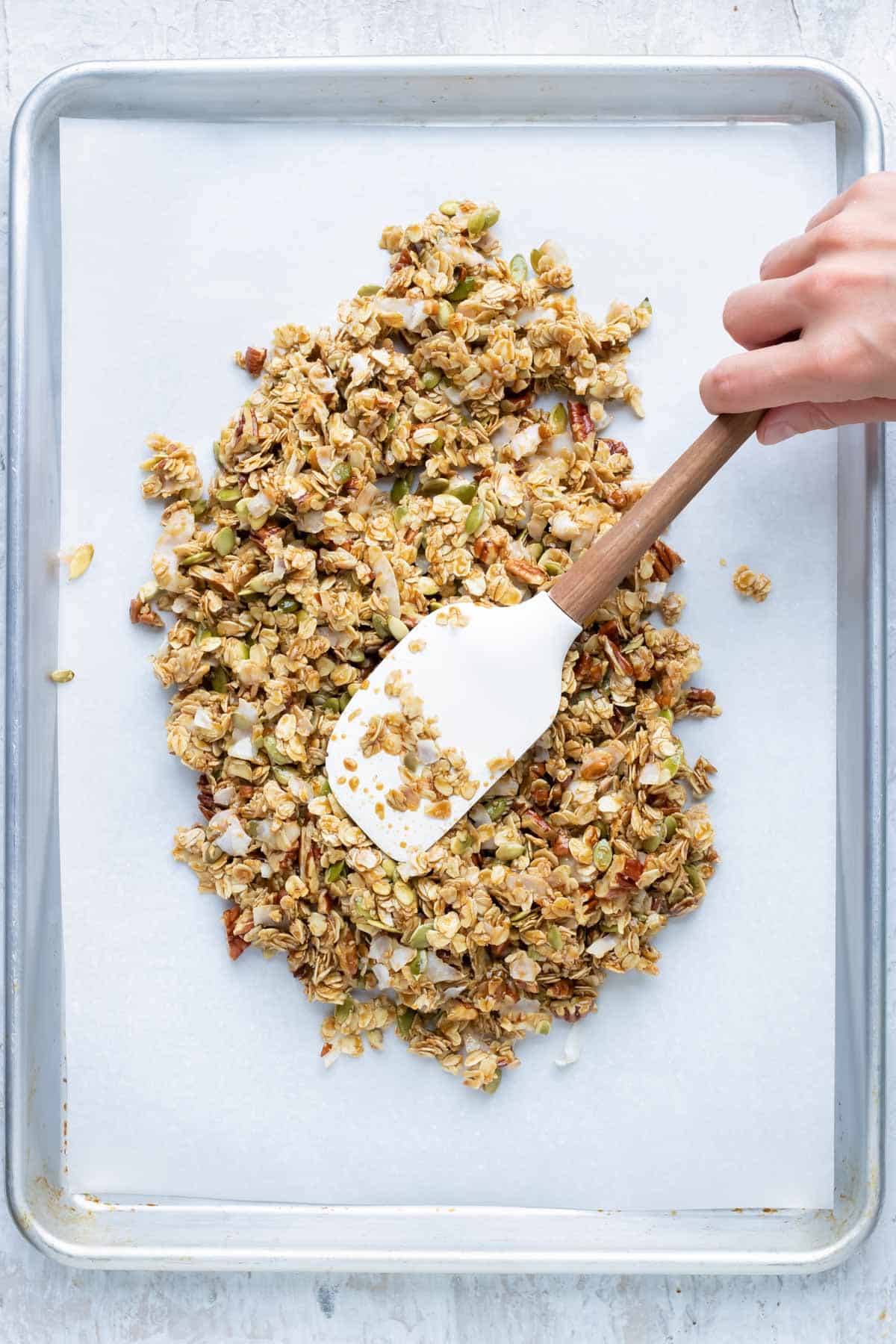 A spatula spreads the granola mixture on a baking sheet.