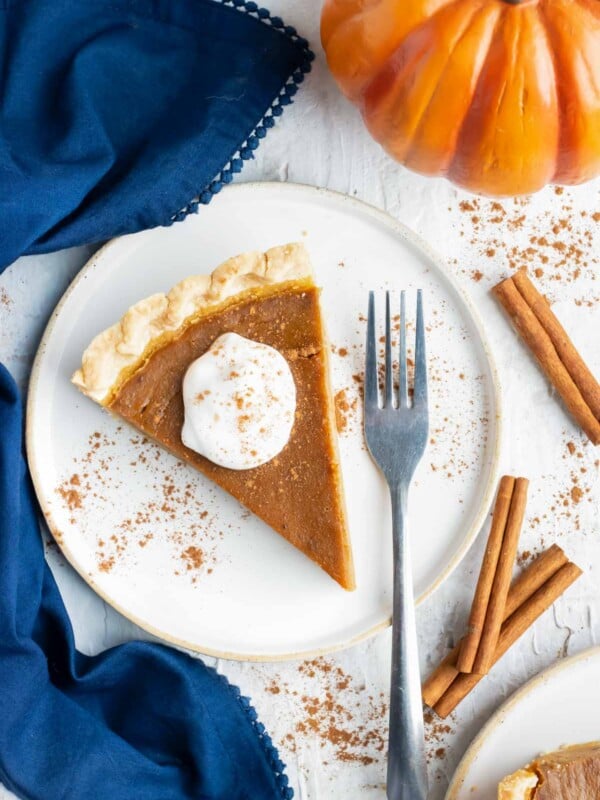 Pumpkin pie slice on a white plate with cinnamon sticks around it.