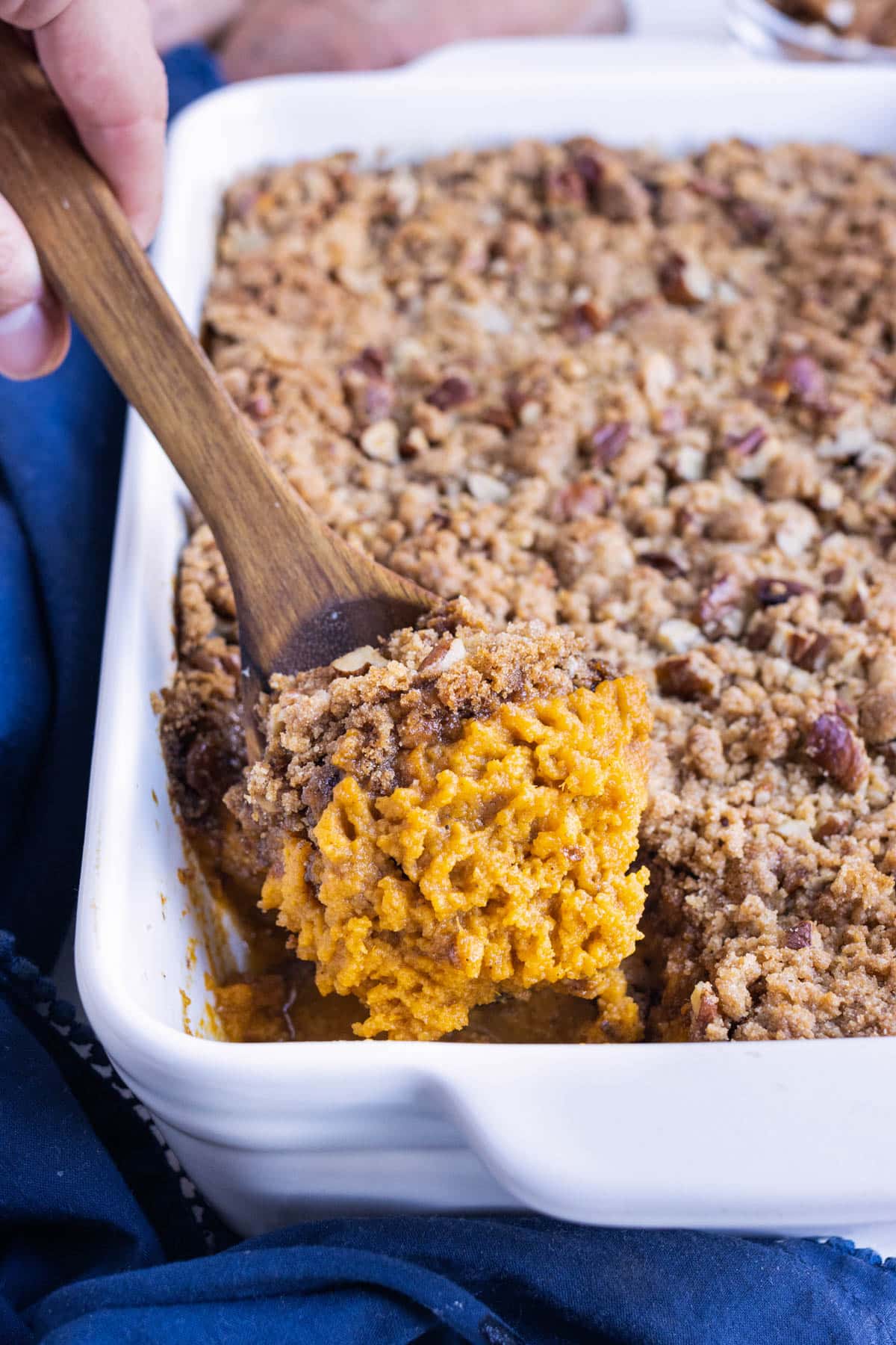 A wooden spoon is scooping up a serving of sweet potato casserole with a brown sugar pecan crumble.