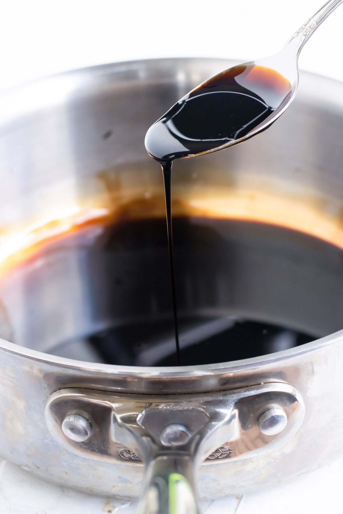 A spoon scooping out a balsamic glaze from a stainless steel saucepan.