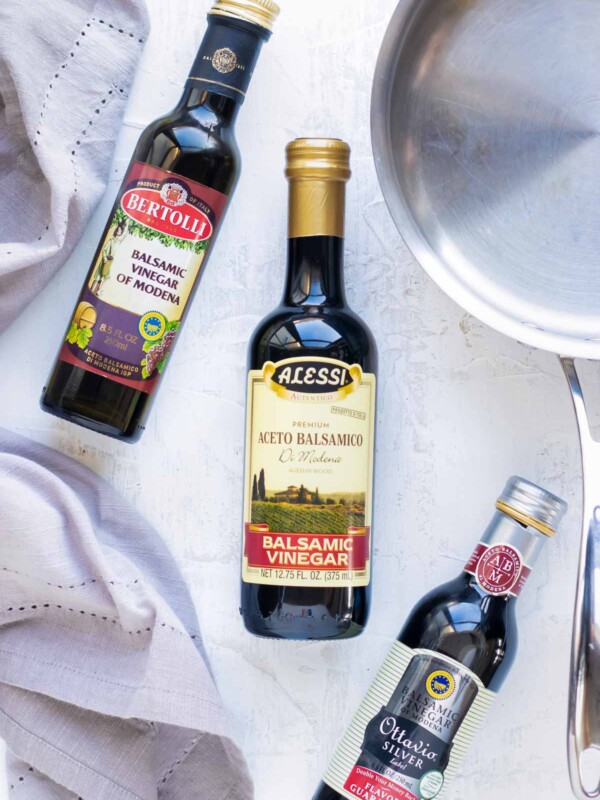 Different brands of balsamic vinegar on a white surface with a stainless steel saucepan.