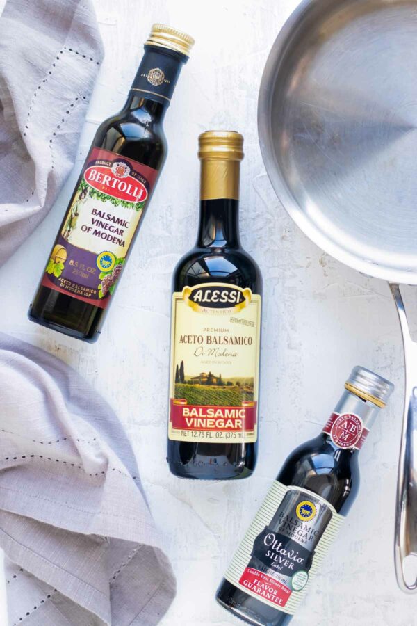 Different brands of balsamic vinegar on a white surface with a stainless steel saucepan.