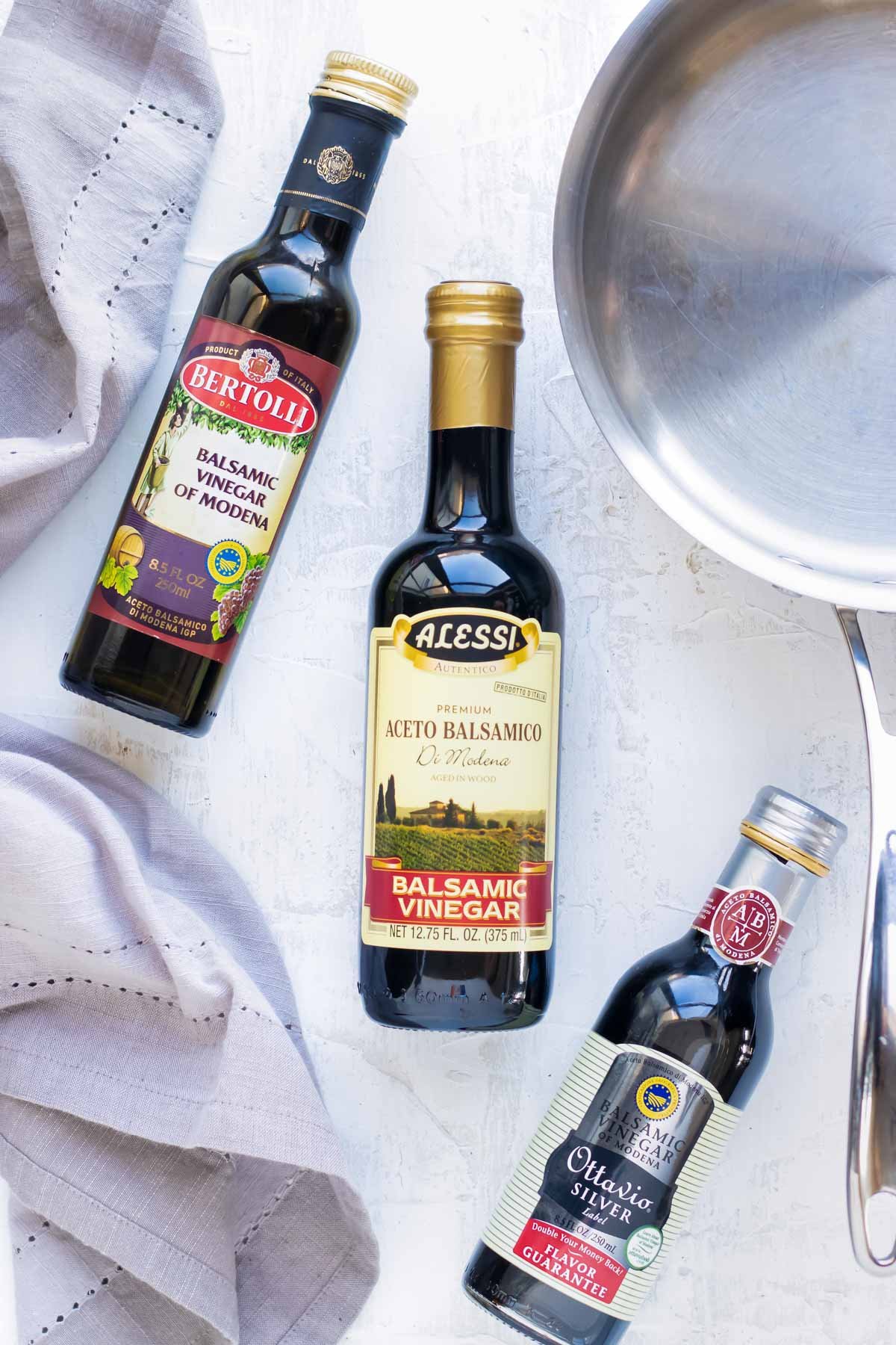 Different brands of balsamic vinegar on a white surface with a stainless steel saucepan to make a balsamic glaze.