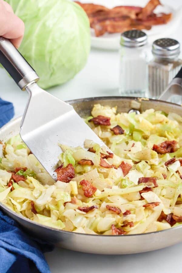 A spatula stirs the cabbage and bacon.