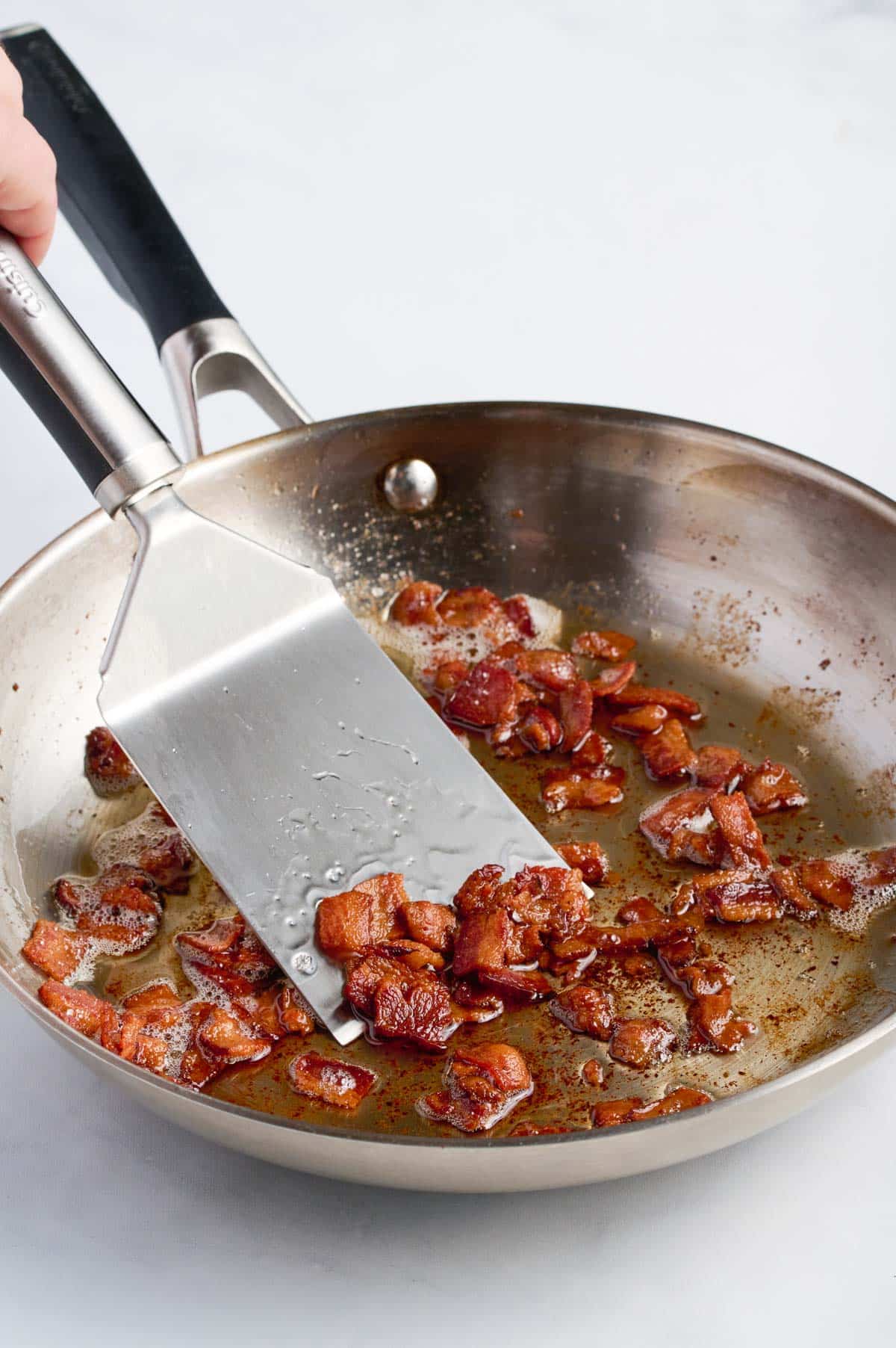 A spatula stirs bacon in a skillet.