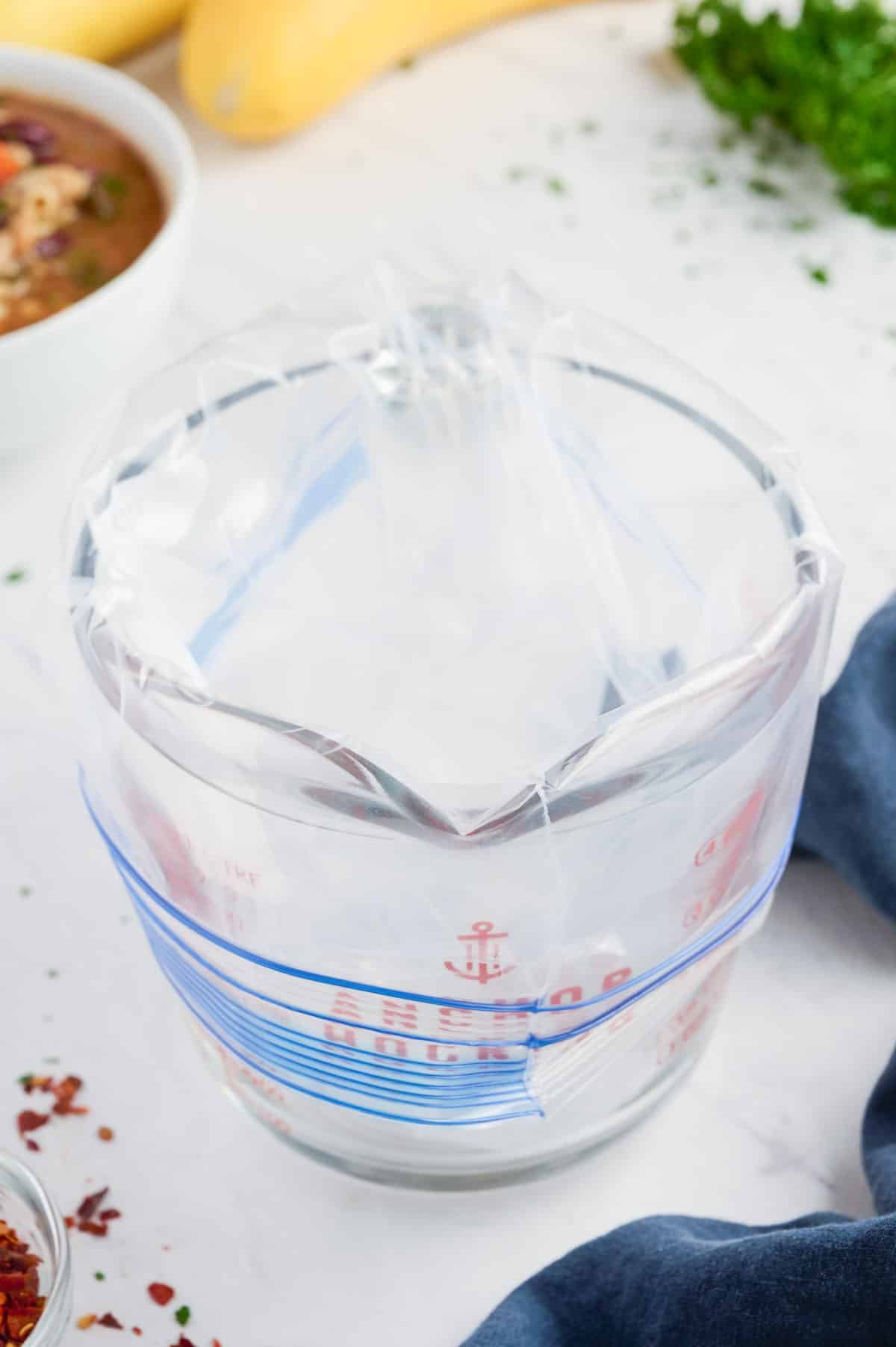 A glass measuring cup with a gallon-sized plastic bag's edges rolled around the top.