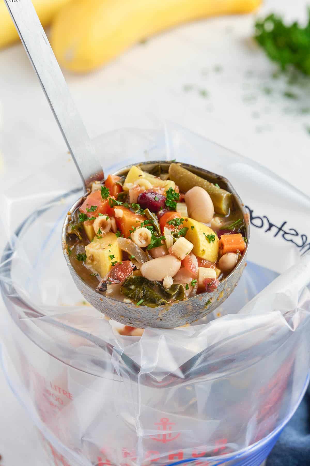 A ladle of minestrone soup entering a gallon-sized plastic bag.