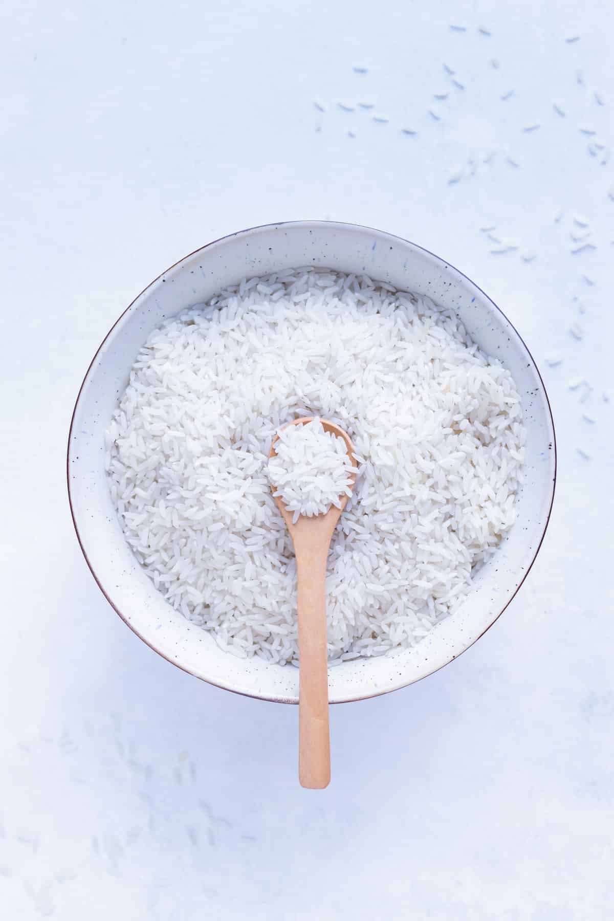 A big, white-speckled bowl full of cooked white rice with a small wooden spoon in the center holding some white rice.