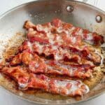 Bacon slices are cooked in a stainless steel skillet.
