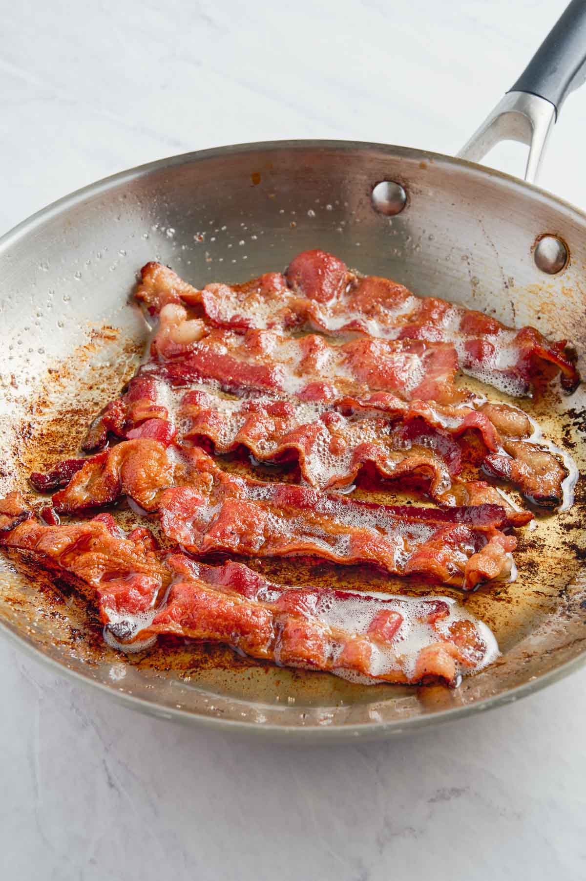 Bacon slices are cooked in a stainless steel skillet.