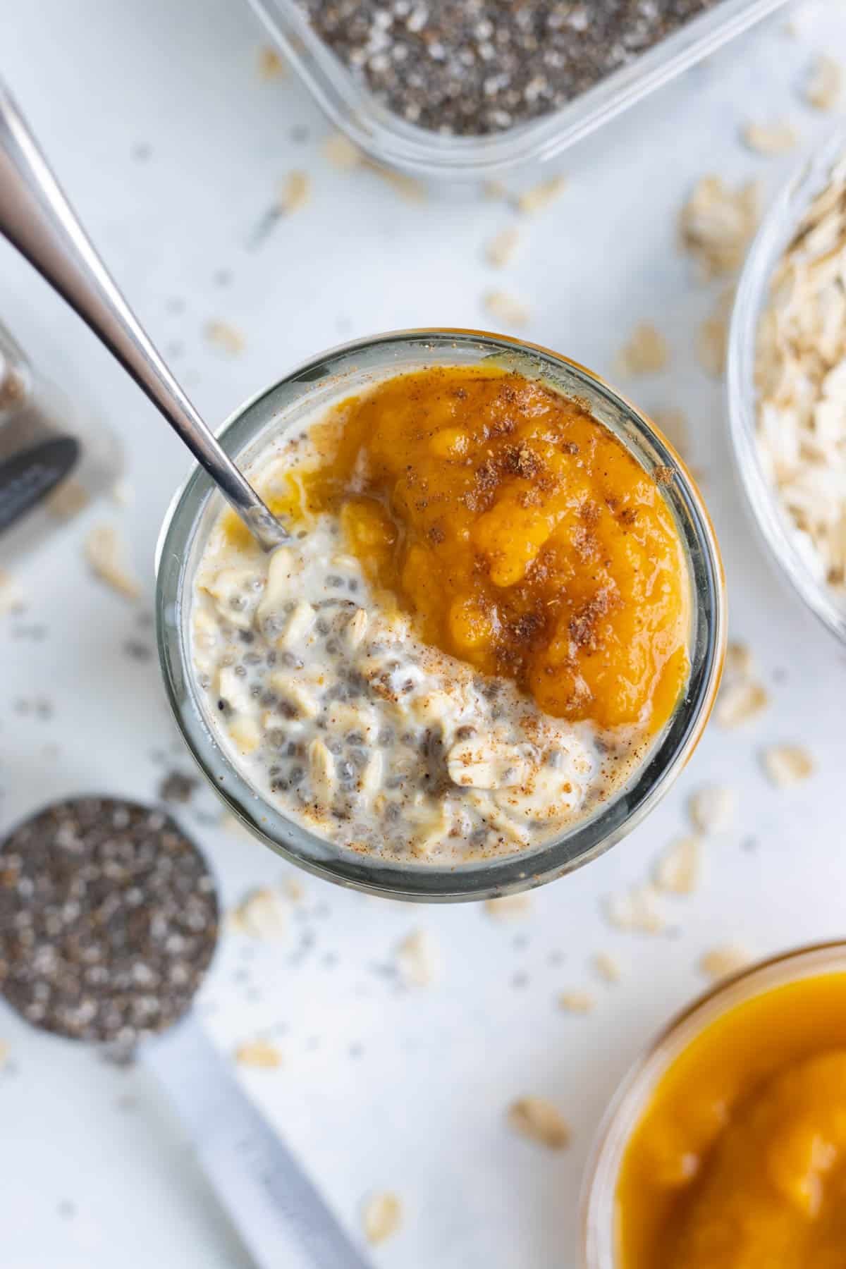 A close-up of a glass mason jar full of overnight oats, pumpkin pie spice puree, toasted pecans, and whipped cream.