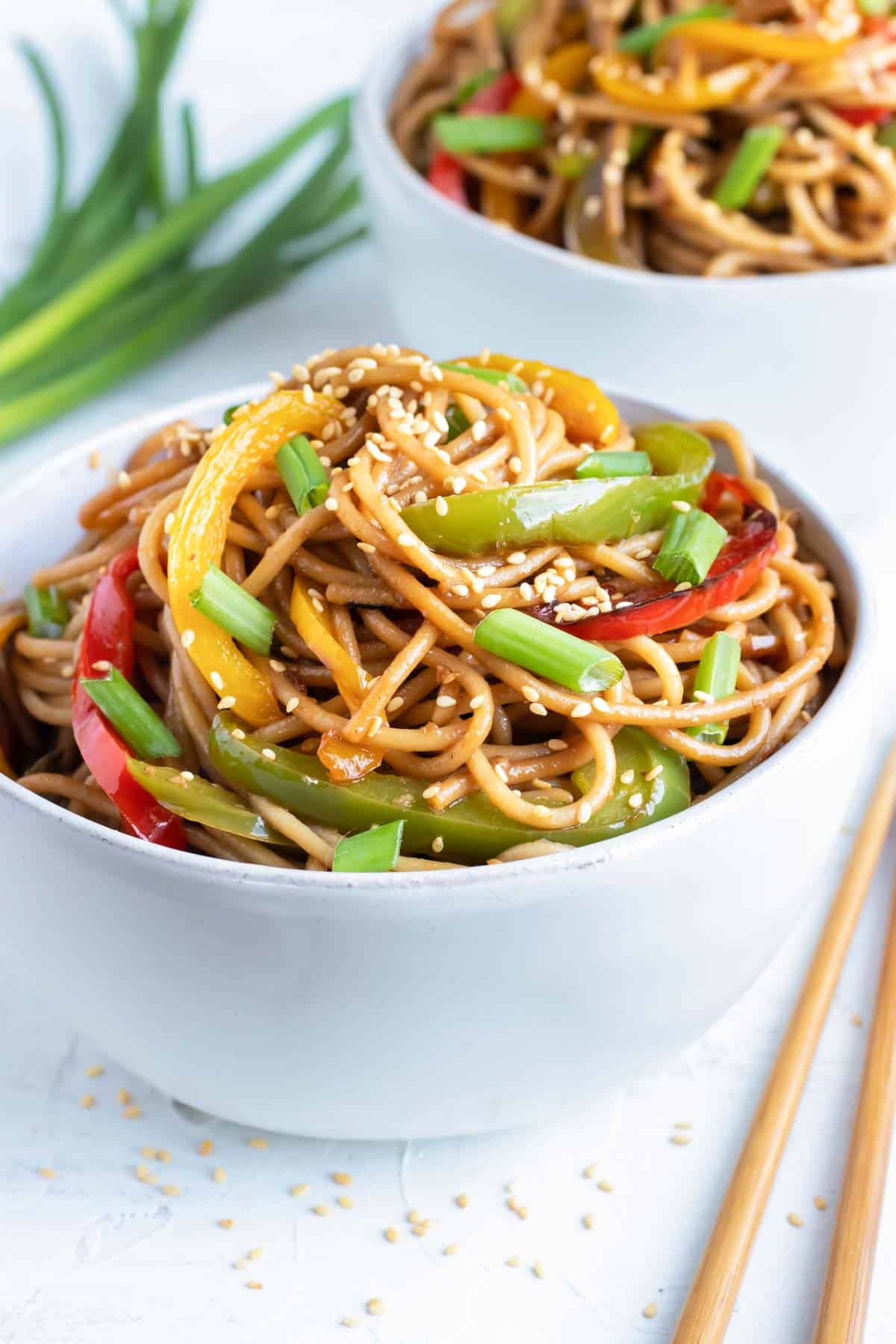Cold sesame noodles with bell peppers in a white bowl with chopsticks and green onions next to it.