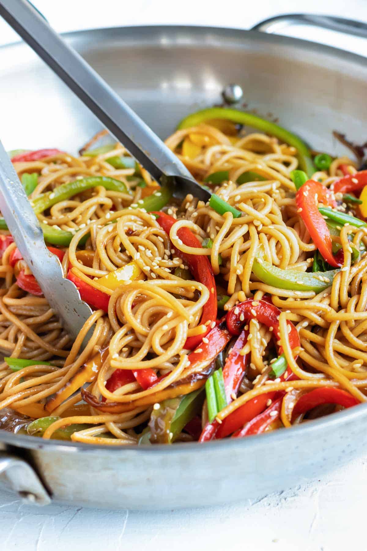 Metal tongs tossing sesame noodles with bell peppers in a stainless steel skillet.