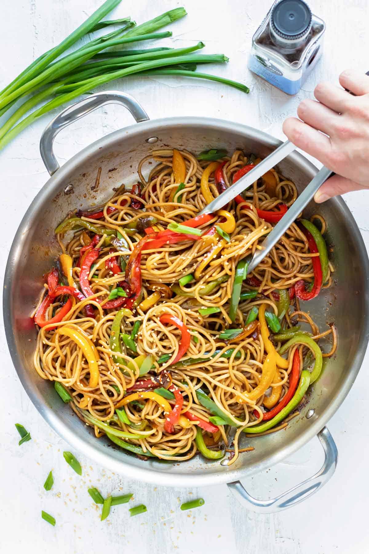 A hand with tongs tossing around a wok full of sesame noodles with garlic, ginger, and bell peppers.
