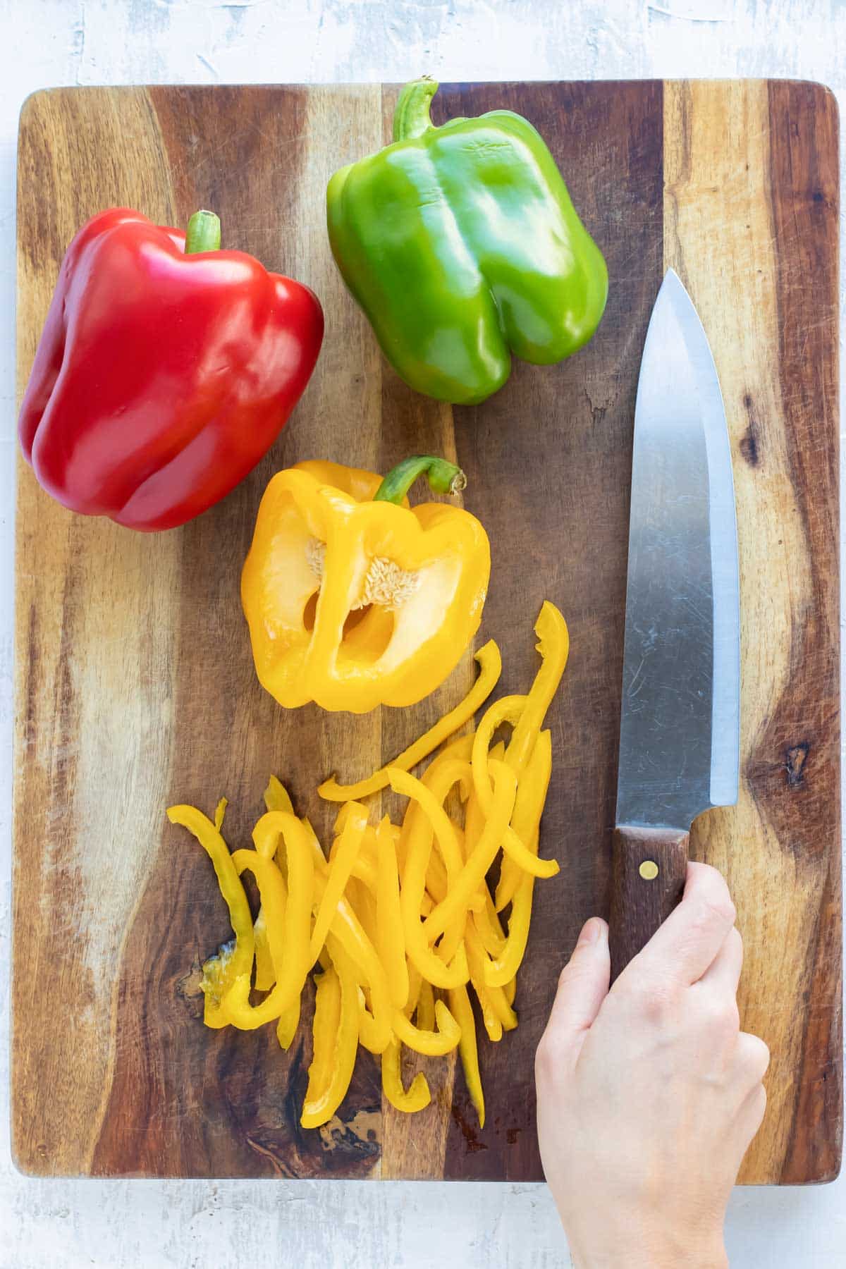 A red, yellow, and green bell pepper on a wooden cutting board being thinly sliced.