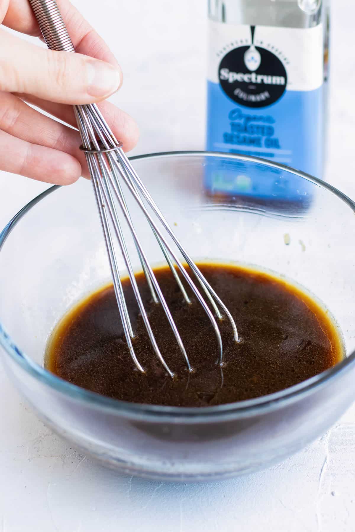 Whisking together the sesame sauce ingredients in a clear bowl with a bottle of toasted sesame oil.