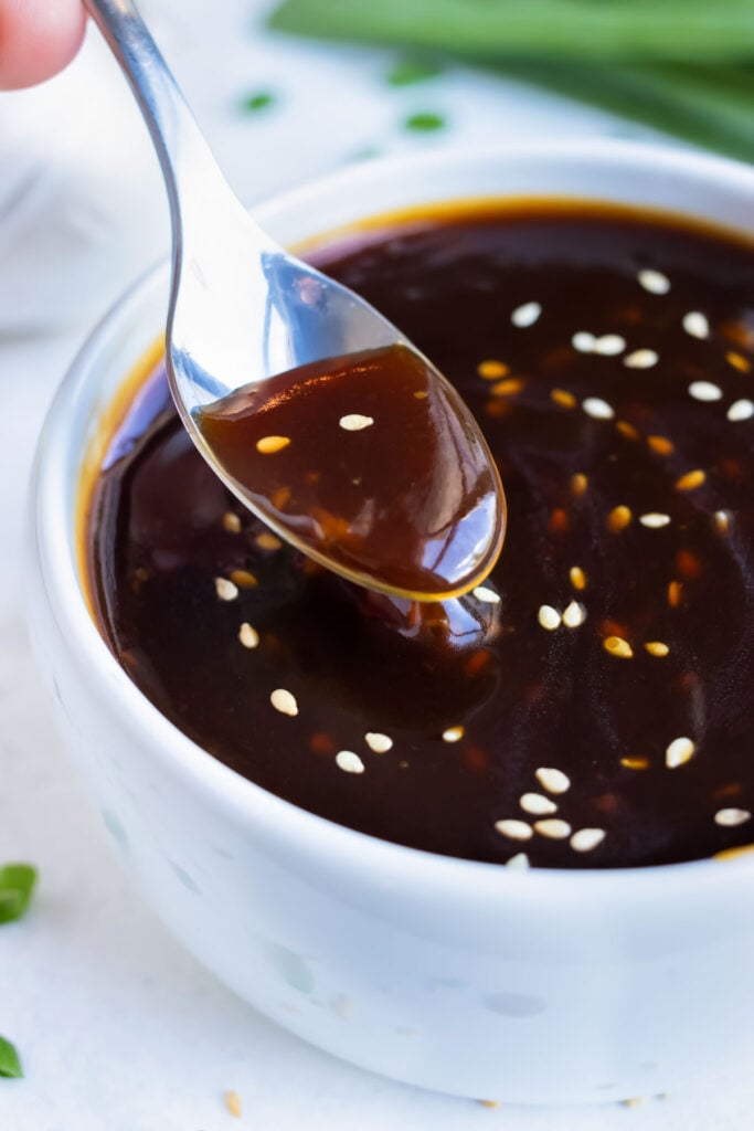A spoon is lifting up a sticky teriyaki sauce from a white bowl.