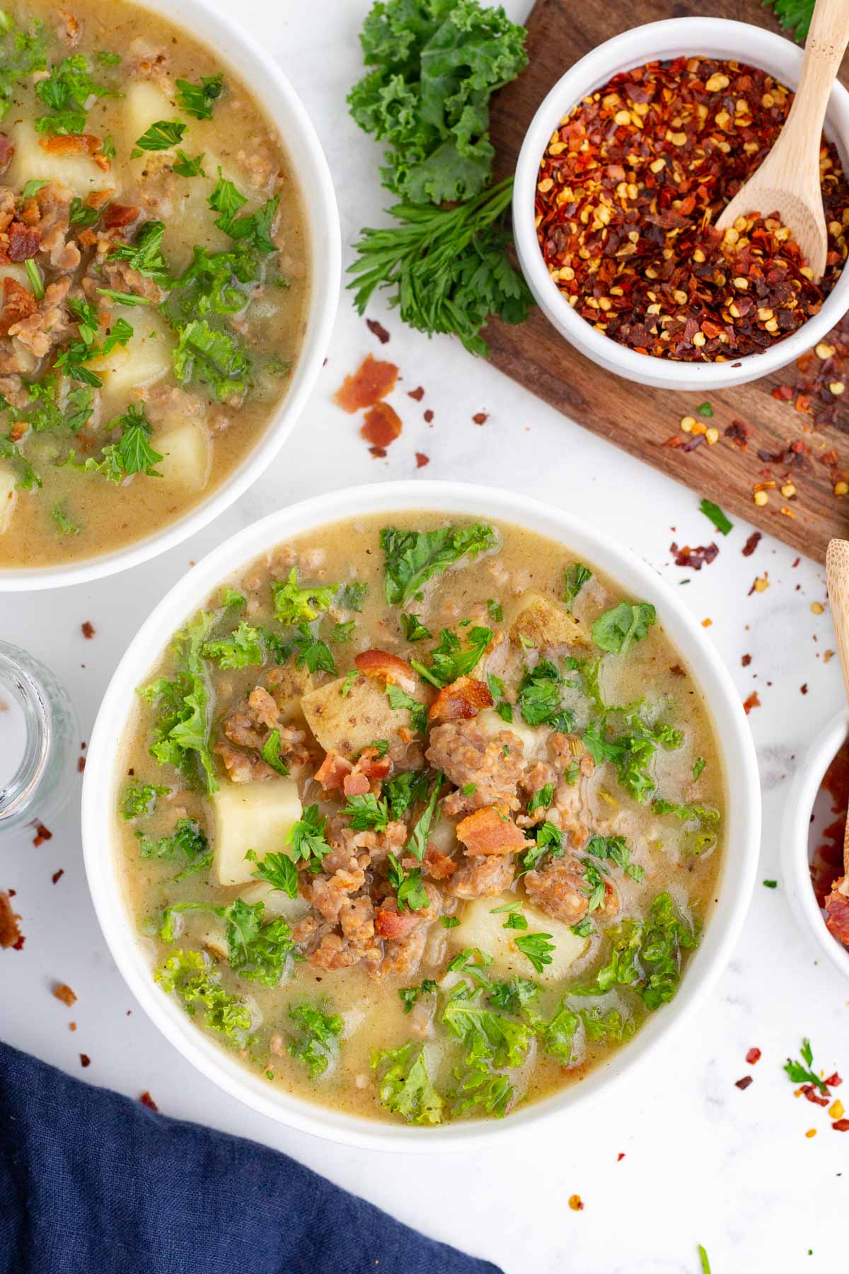 Two white bowls full of a zuppa toscana soup recipe next to kale and red pepper flakes.