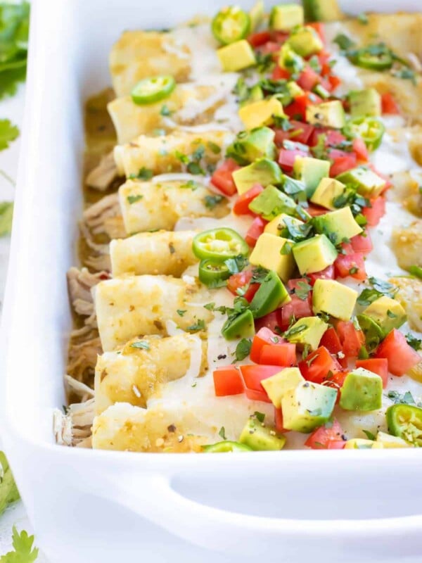 Chicken Enchiladas Verdes in a white baking dish with avocado, tomatoes, and cilantro on top.