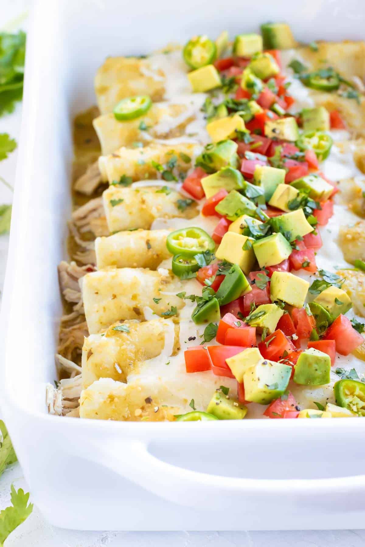 Chicken Enchiladas Verdes in a white baking dish with avocado, tomatoes, and cilantro on top.