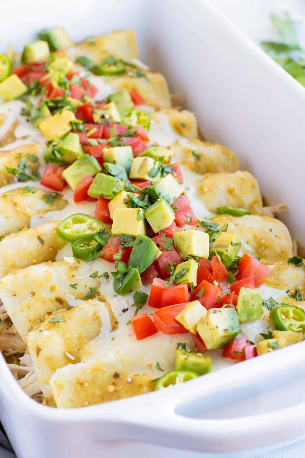 Chicken Enchiladas Verdes recipe in a white baking dish with avocado, tomatoes, and cilantro on top.